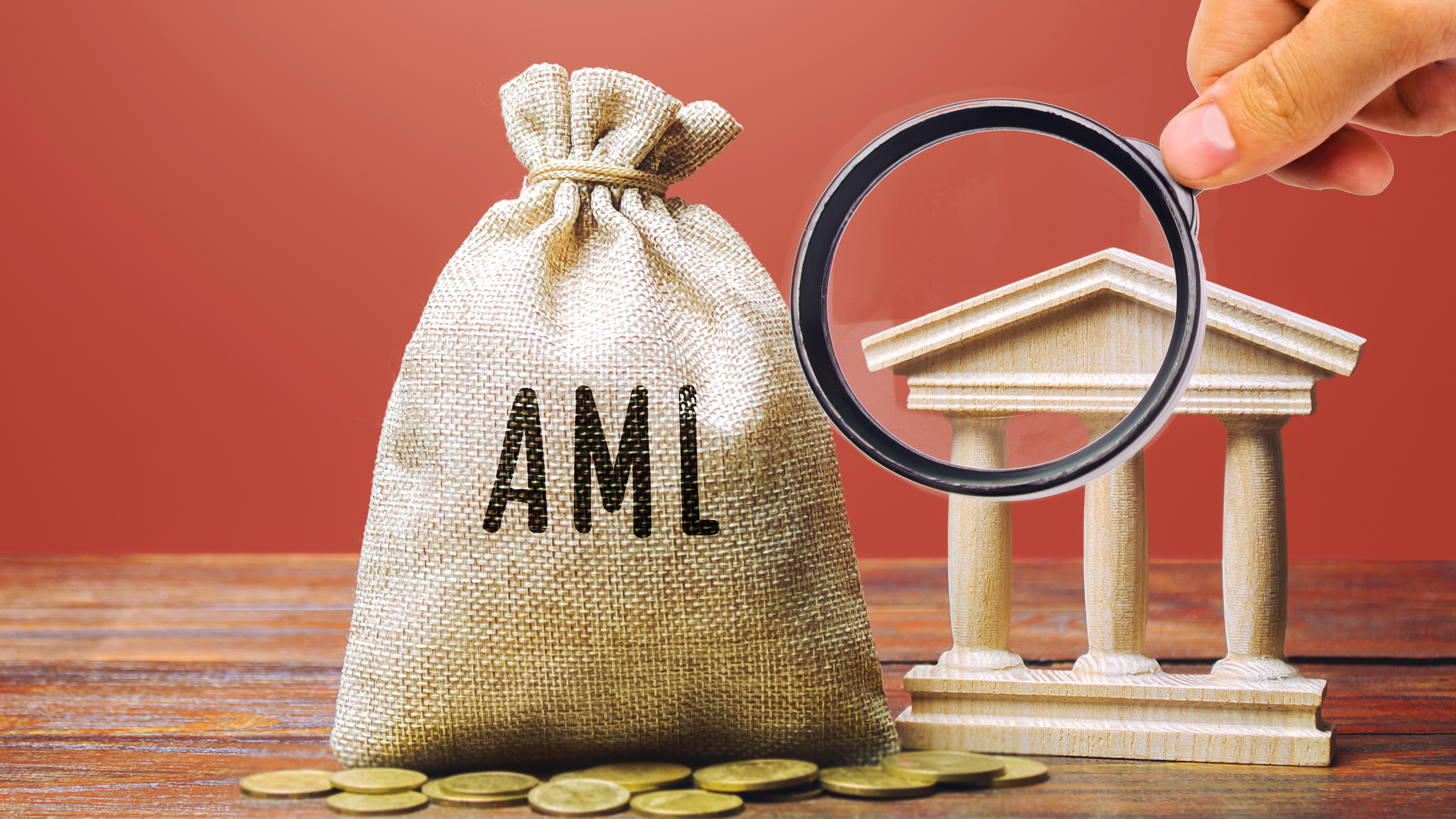A hessian bag with "AML" inscribed on its front, a hand holding a magnifying glass, a model of a bank facade, and coins scattered on a desk. 