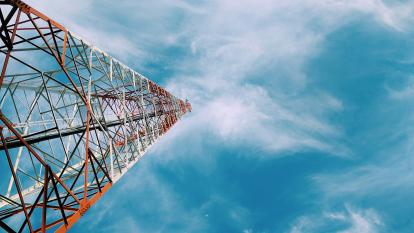 A view of the top of a communications tower from its base, against a blue, clouded sky. 
